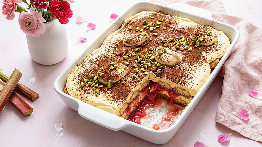 Ein Auflaufdish mit Tiramisu, garniert mit Kakao und gehackten Pistazien. In der Mitte blitzen Rhabarberstücke hervor, die dem Dessert eine fruchtige Note verleihen. Im Hintergrund stehen Blüten in einer Vase, die eine ansprechende Atmosphäre schaffen.