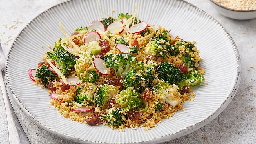 Ein Teller mit einem bunten Salat aus Couscous, Brokkoli, Radieschen und geröstetem Sesam. Der Salat ist frisch und ansprechend angerichtet, mit einer leichten Zitrone garnierte. Die Zutaten sind farbenfroh und verleihen dem Gericht eine gesunde Note.