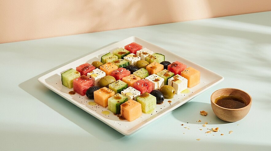 Quadratische Platte mit bunten Würfeln aus Wassermelone, Honigmelone, Gurke und Feta, in Reihen angeordnet, garniert mit schwarzen Oliven und Kräutern; daneben kleine Schale mit Dressing.