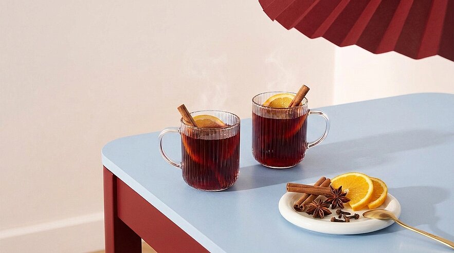 Zwei gläserne Tassen mit dampfendem Glühwein, jeweils garniert mit Orangenscheibe und Zimtstange; daneben ein Teller mit Orangenscheiben, Zimtstangen und Sternanis auf einem Tisch.