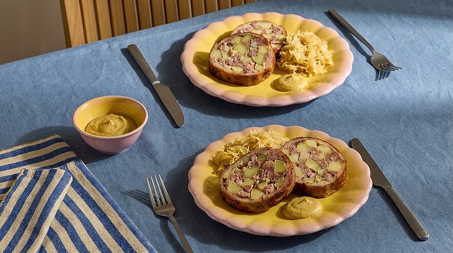 Zwei Teller auf blauem Tuch, jeweils mit zwei Scheiben Fleischterrine in Aspik, Sauerkraut und Senf; Messer und Gabel daneben.