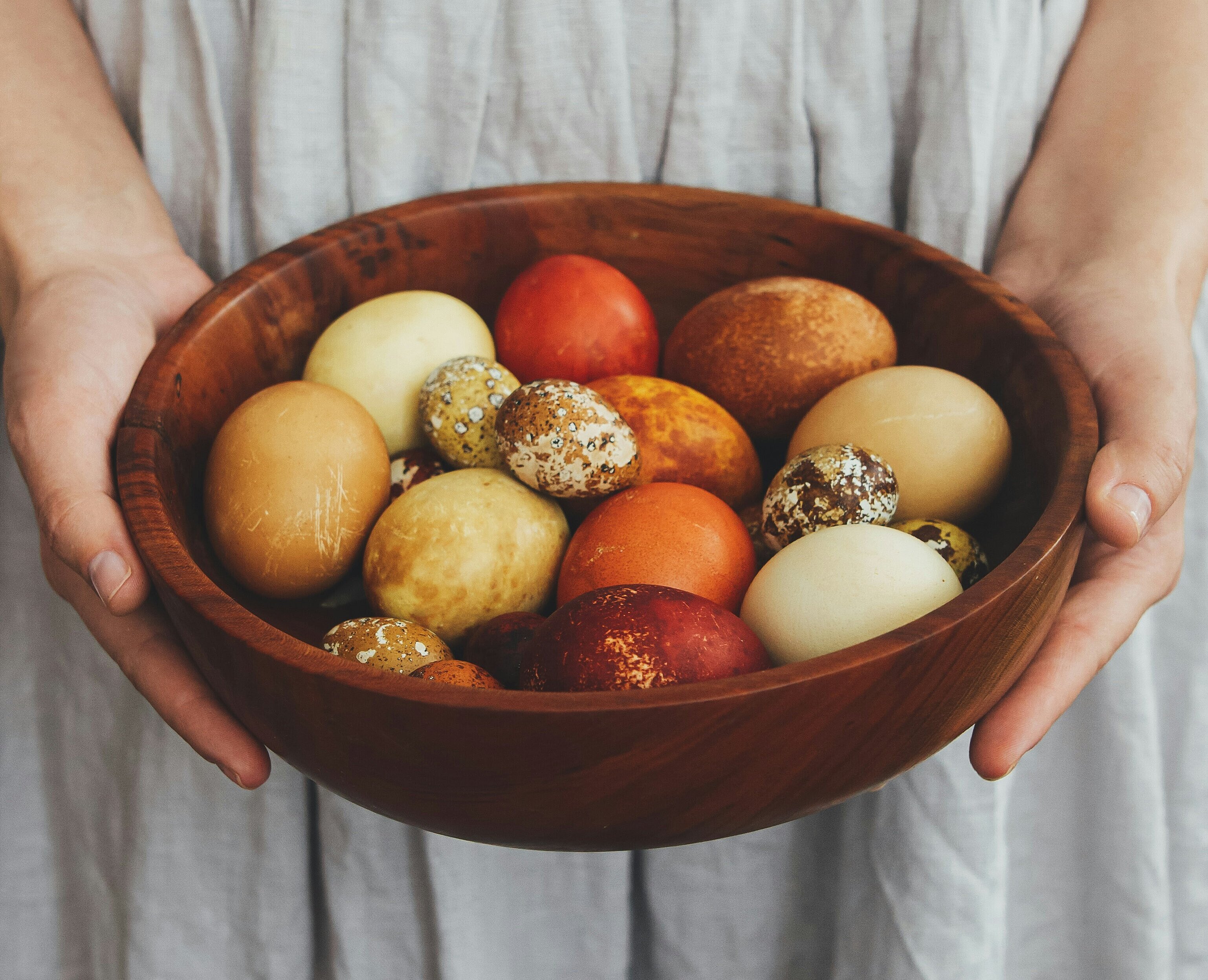 Eine Person hält eine Holzschale mit bunten Eiern, die in verschiedenen Farben und Designs gestaltet sind. Die Eier sind auf einem neutralen Hintergrund angeordnet, während die Person ein einfaches, helles Kleid trägt.