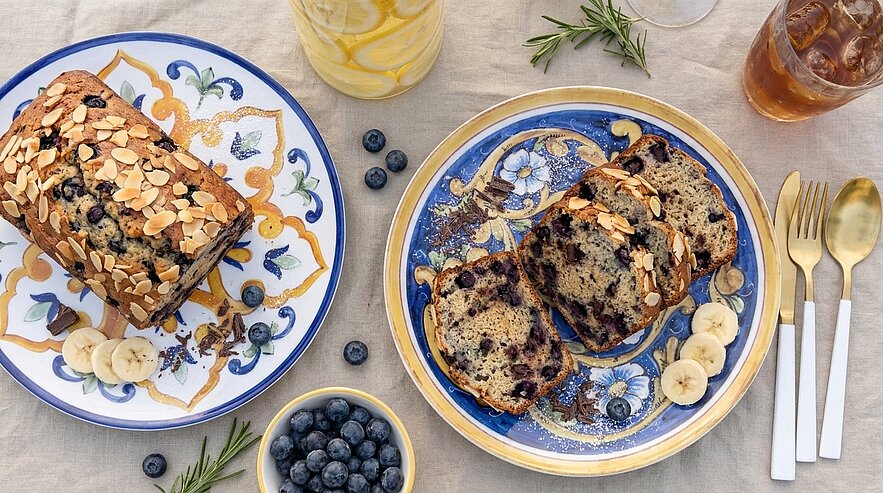 Zwei Bananen-Blaubeer-Kuchen, einer ganz, einer in Scheiben auf dekorativen Tellern, bestreut mit Mandelblättchen und garniert mit Bananenscheiben; daneben eine Schale mit frischen Blaubeeren.