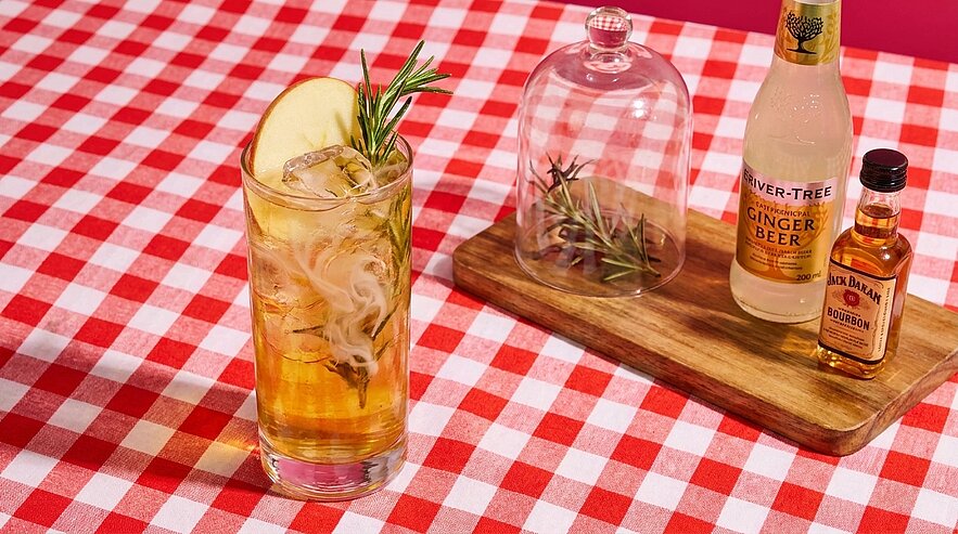 Auf rot-weiß kariertem Tuch steht ein hohes Glas mit hellem Cocktail, Eis, Apfelscheibe und Rosmarinzweig; daneben eine Flasche Ginger Beer und eine kleine Bourbon-Flasche.