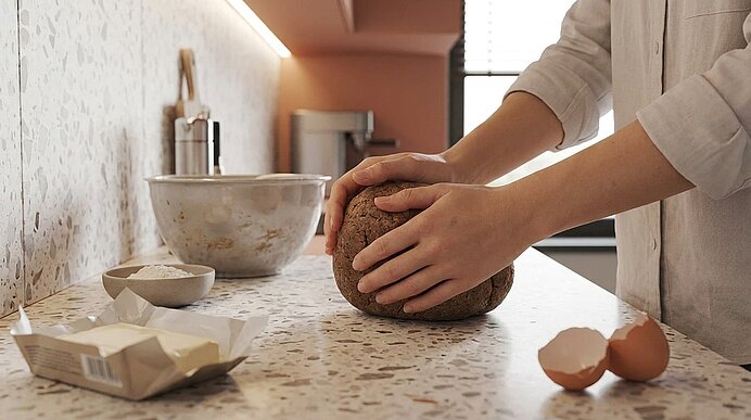 Hände einer Person formen einen runden Brotteig auf der Küchenarbeitsfläche; Butter, Schüssel mit Mehl und eine zerbrochene Eierschale liegen daneben.