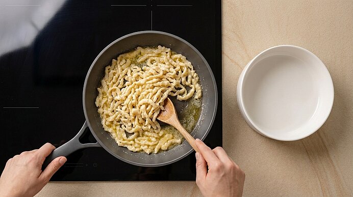 Hände rühren Spätzle in einer Pfanne auf dem Herd mit einem Holzlöffel; daneben eine leere weiße Schüssel.