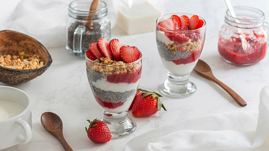 Zwei Gläser mit Schichtdessert sind auf einem weißen Tisch platziert. Jedes Glas enthält Joghurt, Chiasamen, Erdbeermarmelade, Hafergranola und frische Erdbeerscheiben obenauf. Daneben liegen ein kleiner Löffel und einige frische Erdbeeren.