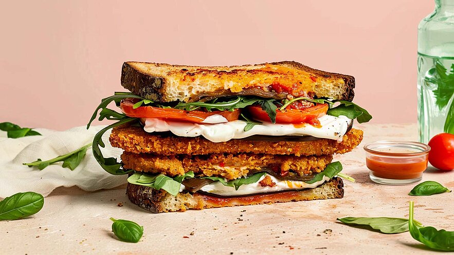 Ein hochgestapelter Sandwich besteht aus knusprigen Zutaten wie paniertem Gemüse, frischen Tomaten und Rucola, mit einer cremigen Sauce zwischen den Schichten. Das Brot ist leicht geröstet und hat eine goldene Farbe. Dahinter ist ein sanfter, rosa Hintergrund zu sehen.