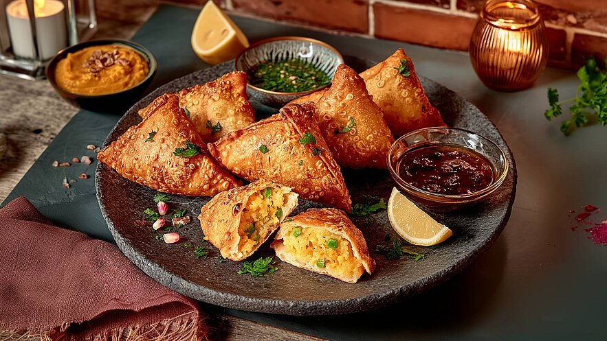 Ein runder Teller mit goldbraunen Samosas, gefüllt mit einer Mischung aus Kartoffeln und Erbsen. Neben den Samosas befinden sich zwei verschiedene Dips sowie eine Zitronenscheibe. Das Gericht ist mit frischem Koriander garniert.