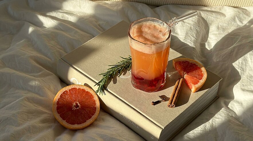 Grapefruitsaft im Glas mit Strohhalm auf einem geschlossenen Buch, daneben halbierte Grapefruit, Rosmarinzweig, Zimtstangen und Nelken auf hellem Bettlaken im warmen Sonnenlicht.