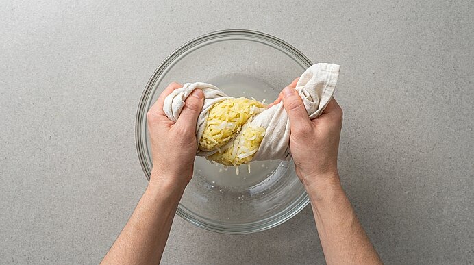 Zwei Hände drücken geriebene Kartoffeln und Zwiebeln in einem Küchentuch über einer Glasschüssel aus.
