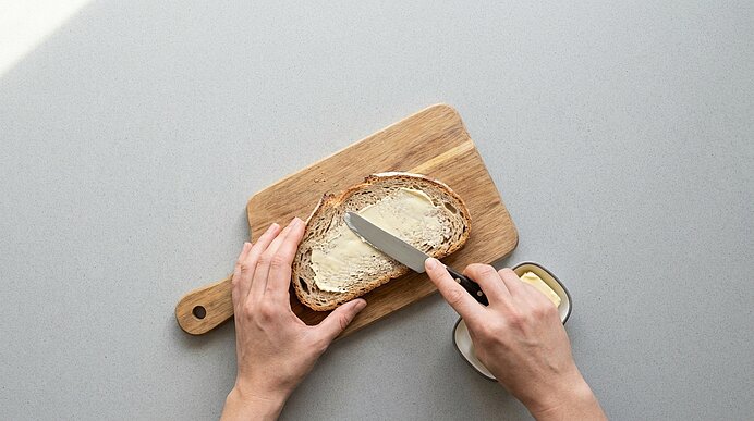 Zwei Hände bestreichen eine Scheibe Vollkornbrot auf einem Holzbrett mit Butter; ein Messer und ein offenes Butterglas stehen daneben.