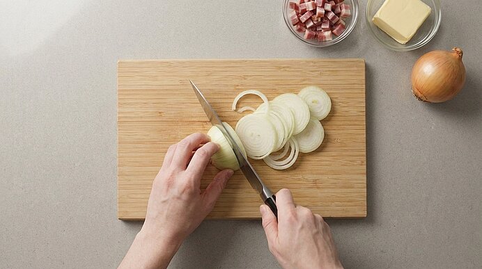 Zwei Hände schneiden eine halbierte Zwiebel auf einem Holzbrett in Ringe; Schüsseln mit Speck und Butter stehen bereit.