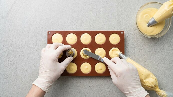 Hände setzen einen gefrorenen Passionsfruchtkern in eine mit Mousse gefüllte Silikonform auf einer Arbeitsplatte.