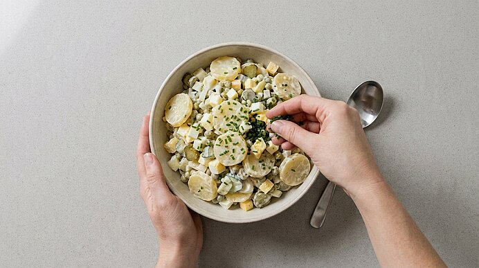 Schüssel mit cremigem Kartoffelsalat; eine Hand hält die Schüssel, die andere streut frisch gehackten Schnittlauch darüber, ein Löffel liegt daneben.