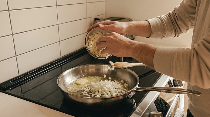 Hände schütten gehackte Zwiebeln aus einer Schüssel in eine Bratpfanne mit schmelzender Butter auf dem Herd.