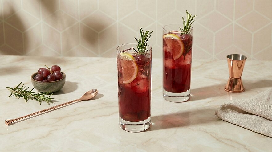 Zwei hohe Gläser mit dunkelrotem Cocktail und Eis, jeweils mit Zitronenscheibe und Rosmarinzweig garniert, auf hellem Marmortisch.