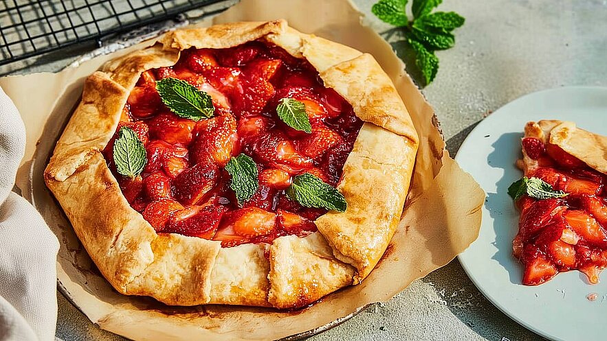Eine frisch gebackene Erdbeer-Galette liegt auf einem Backpapier. Die Füllung besteht aus saftigen Erdbeeren, die mit frischen Minzblättern garniert sind. Ein Stück der Galette befindet sich auf einem Teller daneben, und die krosse Teigkruste ist gut sichtbar.