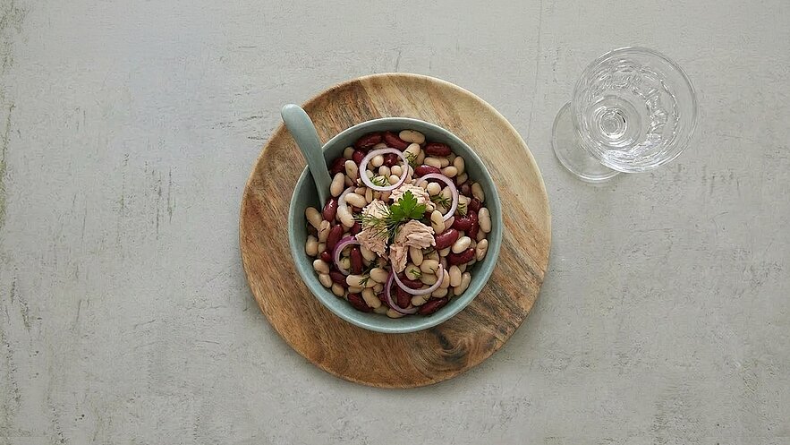 Eine Schüssel mit buntem Bohnensalat, in der sich rote und weiße Bohnen sowie Zwiebelringe und Stücke von Thunfisch befinden. Die Schüssel ist auf einem runden Holzbrett platziert, daneben steht ein Glas Wasser.