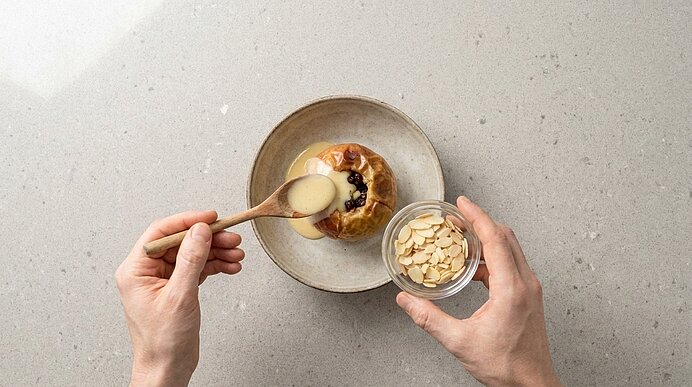 Gebackener Apfel in einer Schüssel; Hände gießen mit einem Löffel Vanillesoße darüber und halten ein Glas mit Mandelblättchen zum Bestreuen.