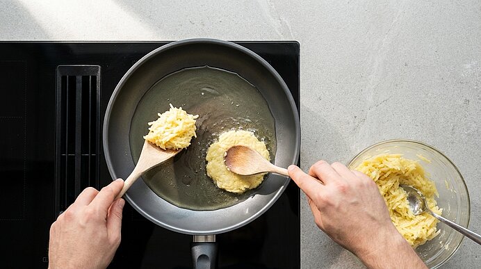 Zwei Hände formen mit Holzlöffeln flache Portionen aus einer Teig- oder Kartoffelmischung und geben sie in eine Pfanne auf dem Herd; rechts eine Schüssel mit Restmasse.