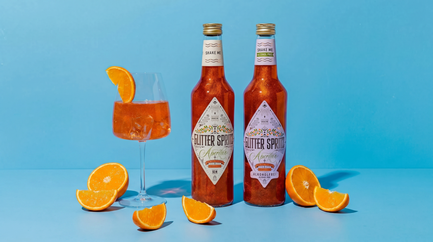 Zwei Flaschen orangefarbener Spritz-Drink neben einem gefüllten Weinglas mit Orangenscheibe und verstreuten Orangenscheiben auf eisblauem Hintergrund.