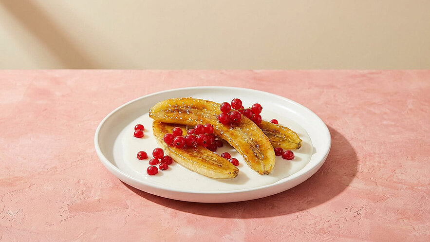 Auf einem weißen Teller liegen gebratene Bananen, die goldbraun sind. Um die Bananen herum sind einige rote Johannisbeeren angeordnet. Der Teller steht auf einer rosa Oberfläche.