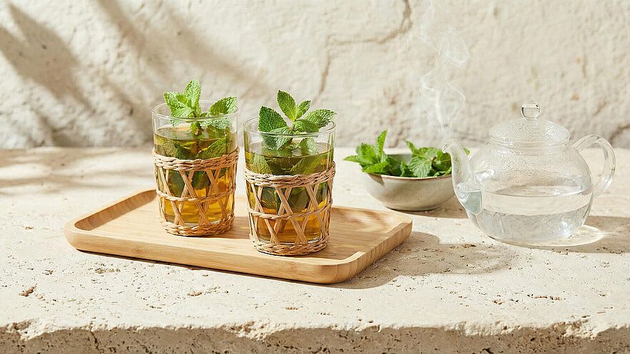 Zwei Gläser mit grüner Teemischung und frischer Minze stehen auf einem Holztablett. Neben den Gläsern liegt eine Glas Teekanne mit heißem Wasser, während auf einem kleinen Teller zusätzliche Minzblätter bereitliegen. Die Szenerie vermittelt ein Gefühl von Frische und Gemütlichkeit.