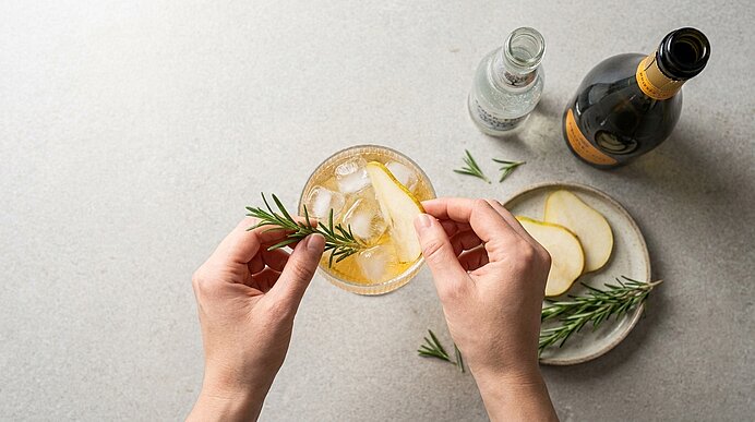 Draufsicht: Hände garnieren ein mit Eis und hellgelbem Getränk gefülltes Cocktailglas mit Rosmarinzweig und Birnenscheibe; daneben Teller mit weiteren Birnenscheiben und Flaschen.