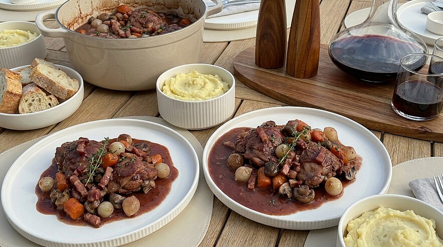 Zwei Teller mit geschmortem Rindfleisch in dunkler Rotweinsauce mit Pilzen, Perlzwiebeln und Karotten, dazu Kartoffelpüree; im Hintergrund ein Schmortopf mit Nachschlag, Brot und eine Karaffe Rotwein.