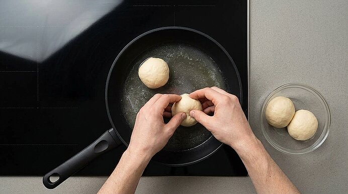 Zwei Hände legen einen Germknödel in eine Pfanne mit Milch-Butter-Mischung auf dem Kochfeld.
