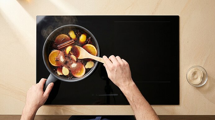 Obenansicht: Hände rühren in einem dampfenden Topf mit Glühwein-Zutaten – Orangenscheiben, Apfelstücke und Zimtstangen – auf einem schwarzen Kochfeld.