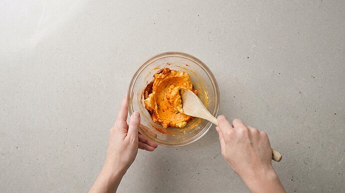 Zwei Hände verrühren Butter und Paprikapulver mit einem Holzlöffel zu einer orangen Masse in einer Glasschüssel.