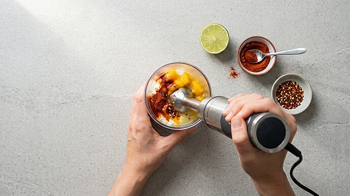 Zwei Hände pürieren mit einem Stabmixer Mangostücke und Gewürze in einem Glas; daneben Limettenscheibe und kleine Schalen mit Chiliflocken und Paprikapulver.