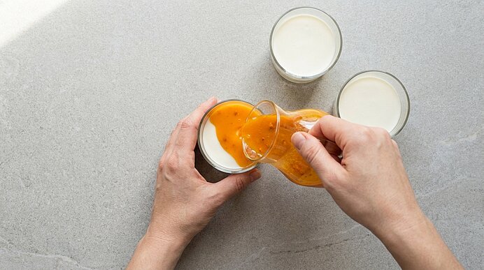 Zwei Hände gießen orangefarbenes Fruchtpüree aus einem kleinen Glas in ein Glas mit weißer Flüssigkeit; zwei weitere Gläser mit weißer Flüssigkeit stehen daneben.