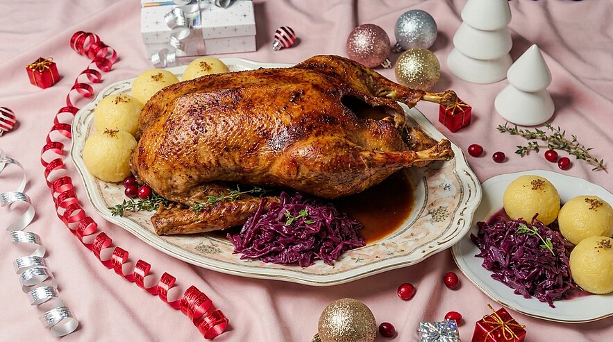 Goldbraun gebratene Gans auf Servierplatte, begleitet von Rotkohl und Kartoffelklößen mit Bratensaft; festliche Dekorationen deuten ein Weihnachtsessen an.