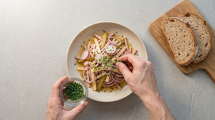 Hände bestreuen eine Schüssel Kartoffelsalat mit gehacktem Schnittlauch; der Salat enthält rote Zwiebelringe und Radieschenscheiben. Daneben liegen Brotscheiben auf einem Schneidebrett.