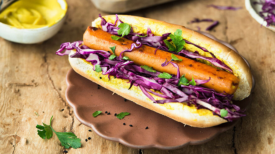 Ein Hotdog auf einem braunen Teller, gefüllt mit einer angegrillten Karotte, lila Krautsalat und frischen Kräutern. Der Hotdog ist mit Senf versehen und ist auf einem Holztisch platziert.