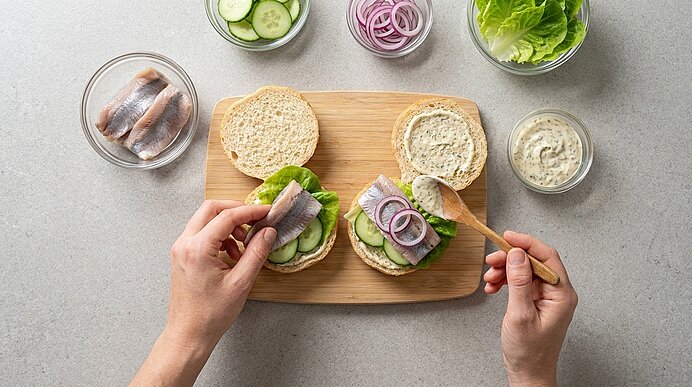 Zwei offene Brötchen auf einem Holzbrett, belegt mit Salat, Gurkenscheiben, eingelegten Heringfilets und roten Zwiebelringen; Hände legen Hering auf das Brot und verstreichen cremigen Aufstrich.