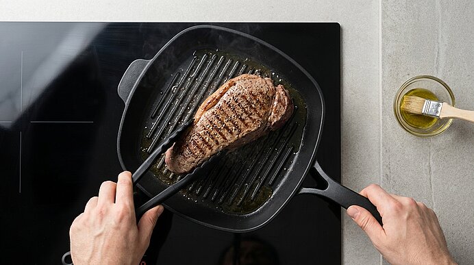 Zwei Hände wenden ein gebratenes Steak mit Grillstreifen in einer Grillpfanne auf dem Herd; eine Hand hält den Pfannengriff. Daneben steht eine kleine Schale mit Öl und einem Pinsel.