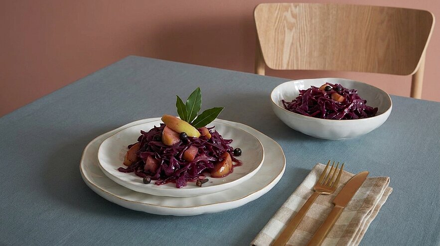 Zwei Servierportionen Rotkohl-Apfel-Salat mit Beeren und Kräutern auf weißem Geschirr, auf einem gedeckten Tisch neben Besteck und Serviette.