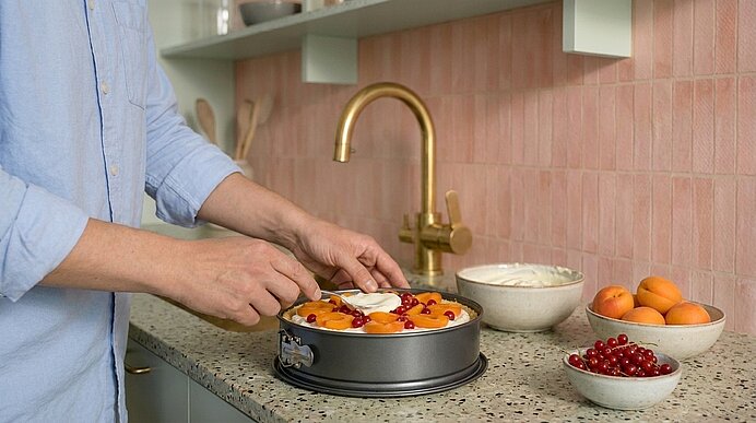 Hände einer Person dekorieren eine Obsttorte in einer Springform auf der Küchenarbeitsplatte; daneben Schalen mit Kirschen, Aprikosen und eine Schüssel Creme.