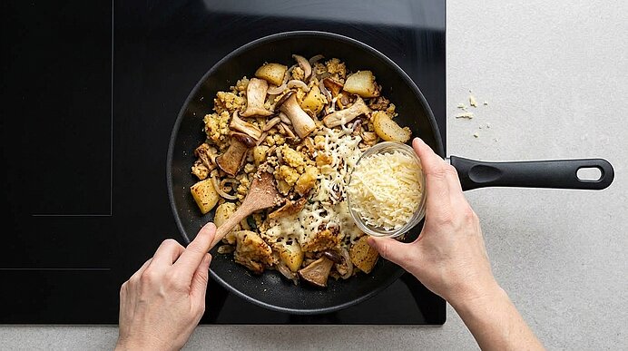 Zwei Hände streuen geriebenen Bergkäse aus einer Schale über den gebratenen Riebelmais mit Pilzen in der Pfanne.