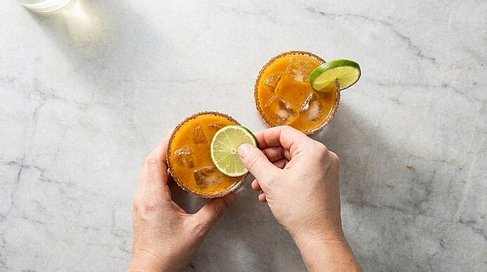 Zwei Hände stecken ein Limettenrad an den Rand eines Glases mit orangefarbener Kürbis-Margarita und Eis.