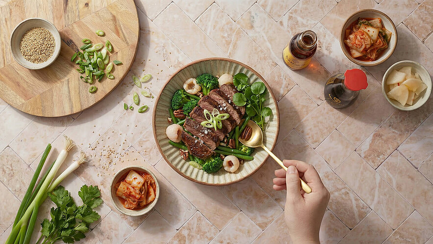 Ein Teller mit aufgeschnittenem Fleisch, umgeben von Brokkoli, grünen Bohnen und Lychee. Eine Hand hält einen Löffel und schöpft das Gericht. In der Nähe sind Schalen mit Kimchi und eingelegtem Gemüse sowie eine Holzplatte mit Sesam und Frühlingszwiebeln.