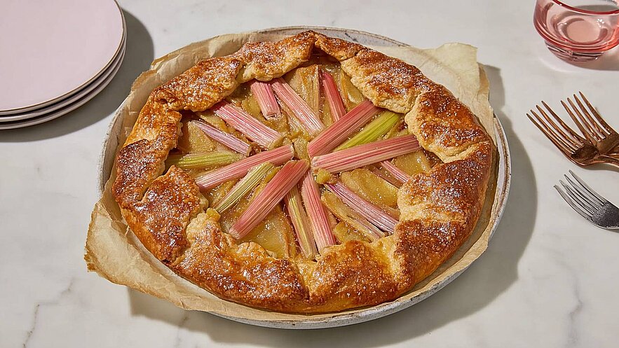 Ein frisch gebackener Rhabarberkuchen in einer runden Form mit goldbrauner Kruste. Der Kuchen ist dekorativ mit Rhabarberstangen angeordnet, die in gleichmäßigen Fächern auf dem Kuchen liegen. Der Hintergrund zeigt eine helle Tischplatte.