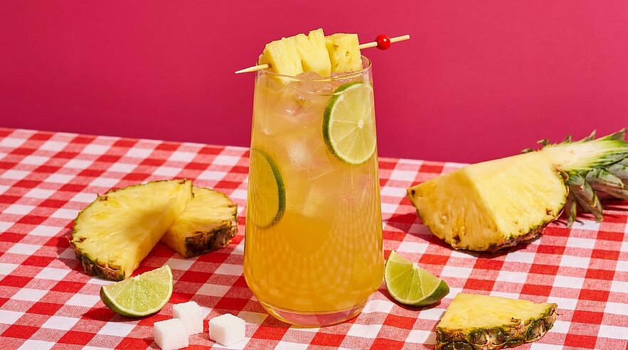 Glas mit eiskaltem Ananas-Limetten-Getränk, Eiswürfeln, Limettenscheiben und Ananasgarnitur auf rot-weiß karierter Tischdecke, umgeben von Ananas- und Limettenspalten.