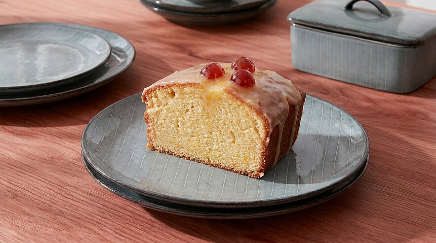 Ein Stück saftiger Kuchen mit Zuckerglasur und Kirschen auf der Oberseite liegt auf einem grauen Teller. Im Hintergrund sind weitere Teller und eine verschlossene Aufbewunsdose zu sehen. Der Tisch ist aus Holz.