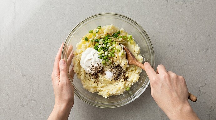 Zwei Hände rühren mit einem Holzlöffel in einer Glasschüssel Kartoffelpüree, verfeinert mit saurer Sahne, gehackten Frühlingszwiebeln, Salz und Pfeffer.
