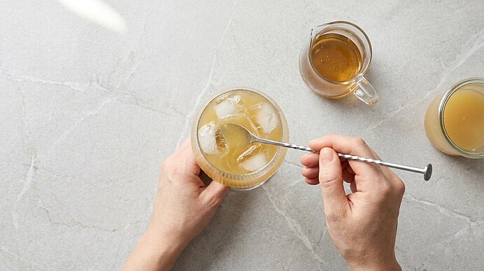Von oben: Hände rühren mit einem langen Barlöffel ein Getränk mit Eis in einem Glas; daneben zwei kleine Kännchen mit brauner und heller Flüssigkeit.
